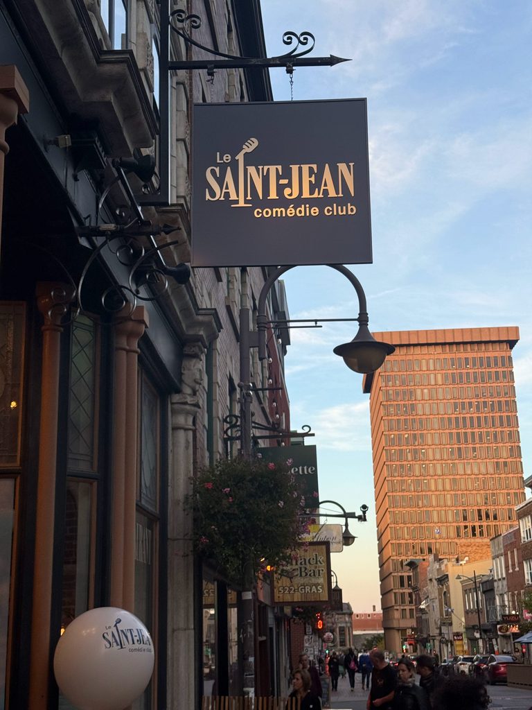 Enseigne du Saint-Jean Comédie Club dans une rue du centre-ville de Québec, avec un bâtiment historique et une tour à bureaux en arrière-plan.